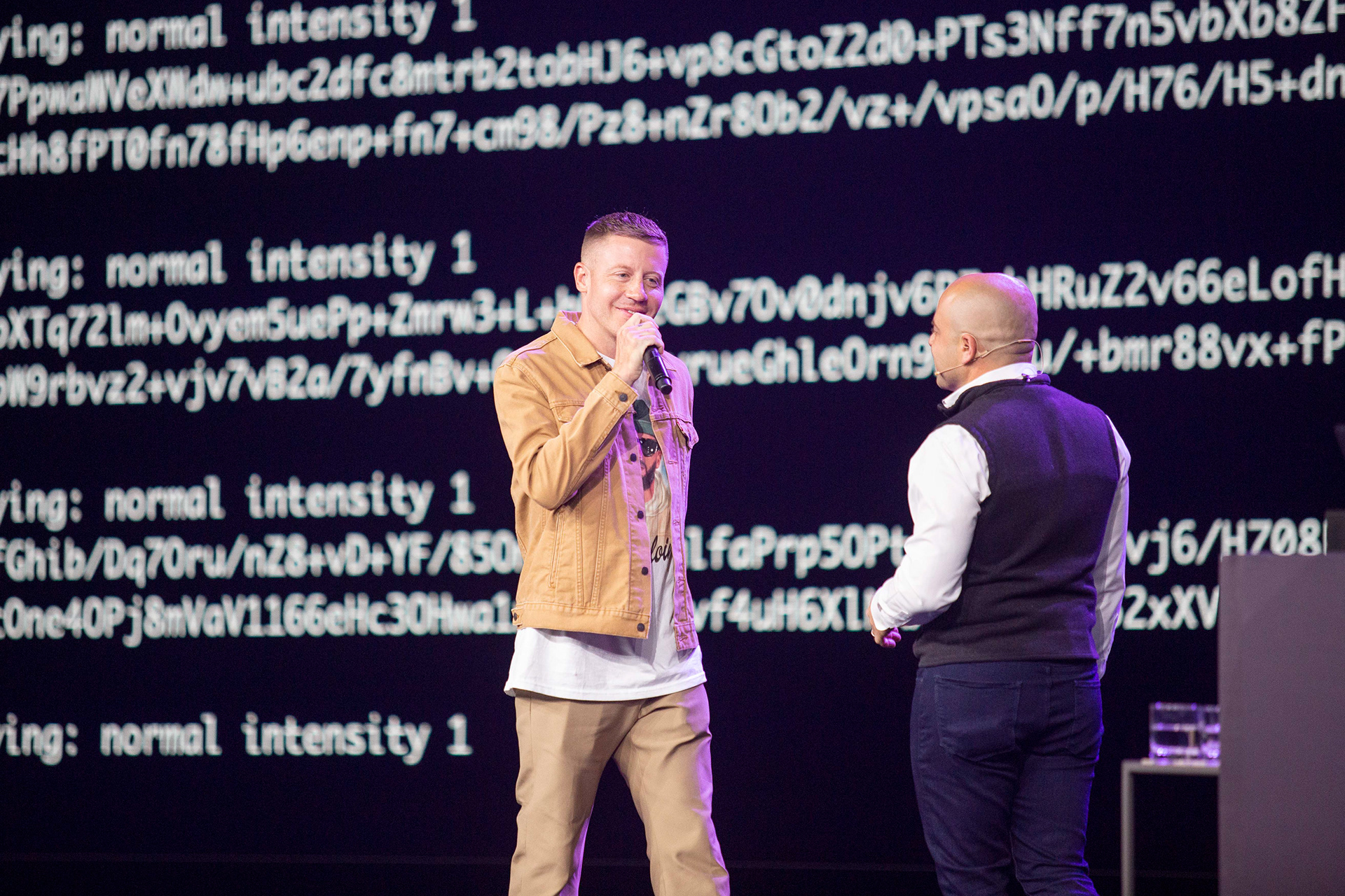 Two speakers on stage with scrolling code displayed on large backdrop